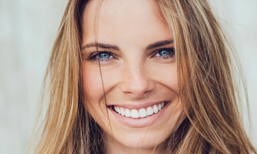 Close-up picture of a smiling happy woman with long brown hair and perfect teeth,  looking directly at the camera and showing her happiness with her All-on-Eight implant-supported denture  she had made at Premier Holistic Dental in beautiful Costa Rica.