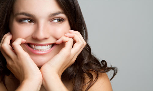Close-up picture of a smiling young woman with long brown hair, holding both hands to the sides of her face, happy with her Holistic dental whitening treatment at Premier Holistic Dental in Costa Rica.