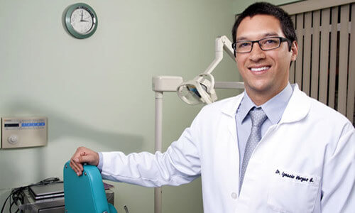 Picture of a smiling Premier Holistic Dental dentist in San José, Costa Rica.  The dentist is wearing a white coat and his arm Is resting on a modern dentist chair.