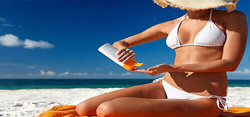 Picture of an attractive woman applying suntan lotion on a beautiful Costa Rica beach while having dental work at Premier Holistic Dental in Costa Rica.  The woman has on a two-piece white bathing suit and she is sitting on a beach towel with a beautiful blue Costa Rica sky in the background.